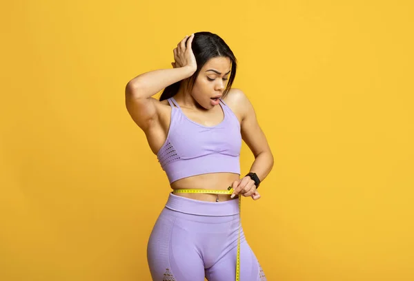 衝撃的な若い黒の女性は、腰にテープメジャーを使用して重量を制御します,頭の上に手を握ります,黄色の背景 — ストック写真