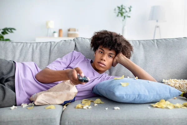 Tembel zenci genç adam kanepeye yayılmış aperatiflerle uzanıyor, uzaktan kumandayı tutuyor, evde televizyon seyrediyor. — Stok fotoğraf