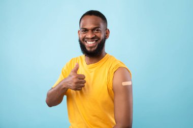 Neşeli Afro adam baş parmak hareketi yapıyor, Coronavirus aşısı aşısından sonra koluna yara bandı yapıştırıyor.