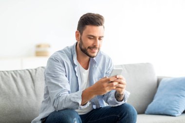 Kullanışlı uygulama konsepti. Milenyum adamı oturma odasındaki koltukta otururken modern akıllı telefonu kullanıyor.