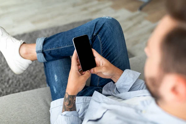 Mockup kép felismerhetetlen fiatal srác használja okostelefon fekete képernyővel, ül a kanapén otthon — Stock Fotó