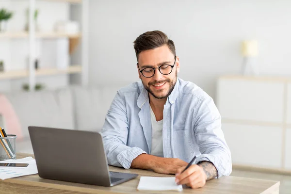 Glücklicher Millennial-Mann, der online nach Arbeit sucht, am Tisch sitzt, Laptop benutzt und Notizen macht, freier Platz — Stockfoto
