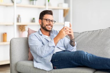 Harika bir mobil uygulama. Mutlu adam akıllı telefon kullanıyor, sosyal ağlarda mesajlaşıyor ve sörf yapıyor, kanepede oturuyor, yer kopyalıyor.