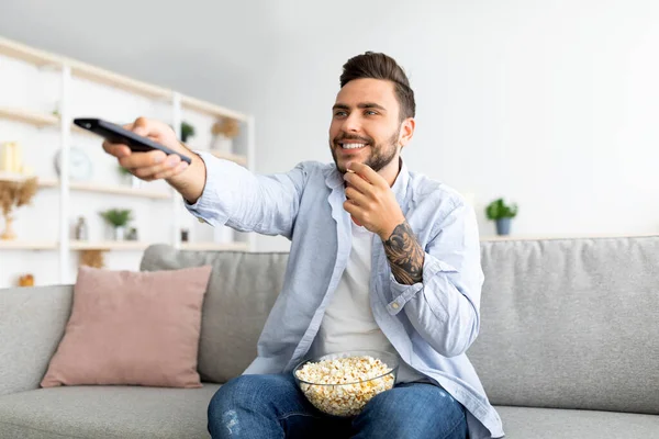 Vrolijke duizendjarige man tv kijken en popcorn eten, van kanaal wisselen met afstandsbediening — Stockfoto