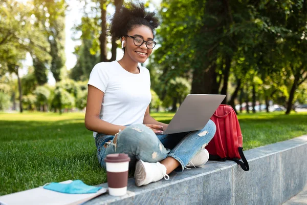 Heureuse étudiante noire utilisant un ordinateur portable portant des écouteurs assis à l'extérieur — Photo