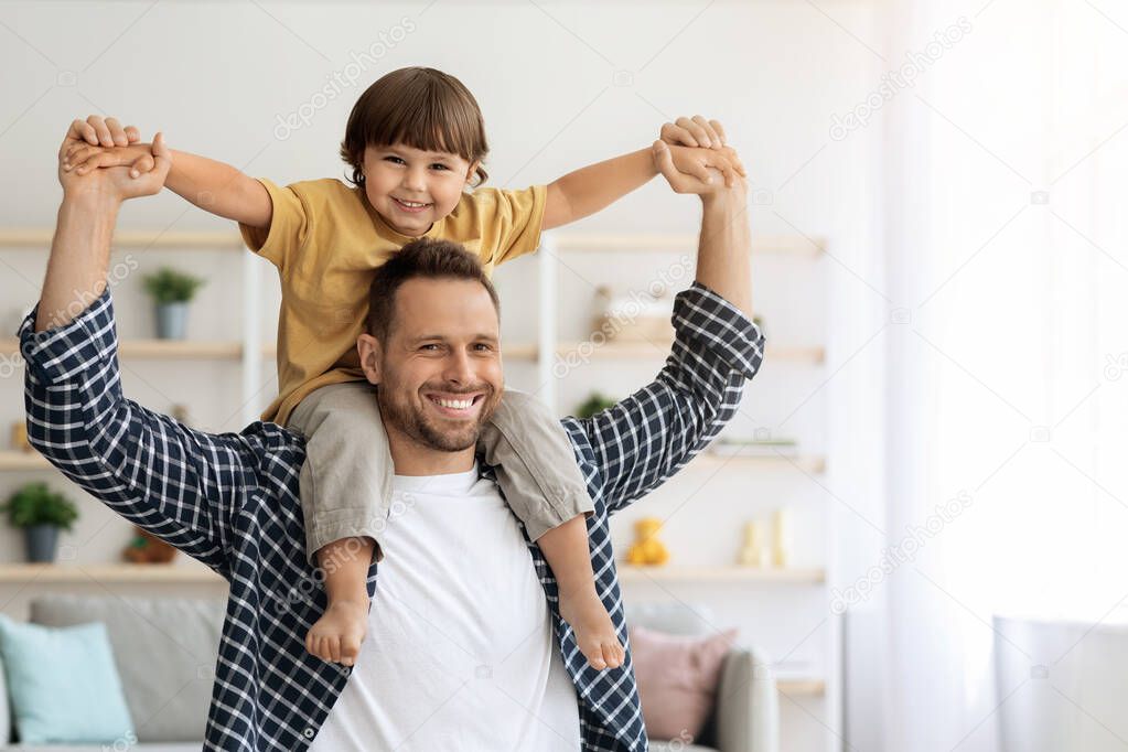 Cheerful father riding his little son on shoulders, having fun together at home, both smiling to camera, empty space