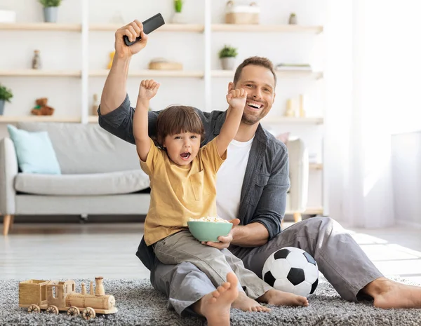 Emocional niño animando al equipo de fútbol favorito, viendo deportes junto con papá, sentado en el suelo en casa — Foto de Stock