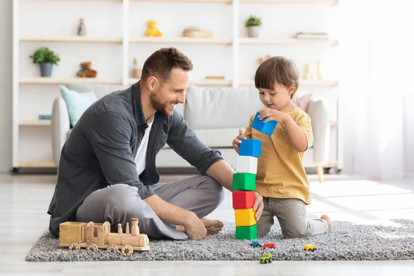 Speeltijd. Vrolijke liefhebbende vader spelen kleurrijke blokken met zijn kleine zoon, jongen bouwen grote toren thuis interieur — Stockfoto