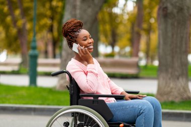 Tekerlekli sandalyedeki güzel siyahi bayan sonbaharda şehir parkında akıllı telefondan konuşuyor.