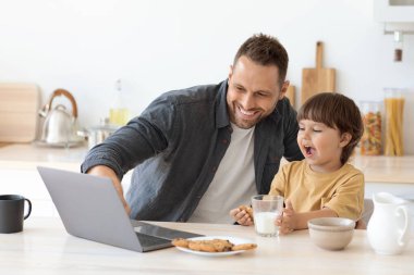 Küçük oğlu mutfakta oturup kahvaltıda laptopla ağ kurarken mutlu genç bir baba.