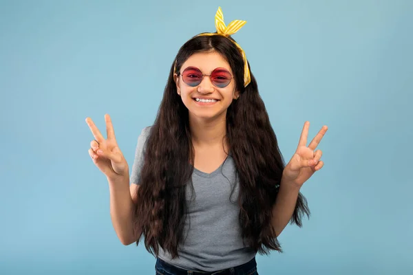Portrait d'une adolescente indienne en lunettes de soleil montrant la paix, la victoire ou deux signes, regardant la caméra sur fond bleu — Photo