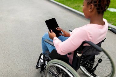 Tekerlekli sandalyedeki genç siyahi kadın sonbahar parkında boş ekranlı tablet bilgisayar kullanıyor.