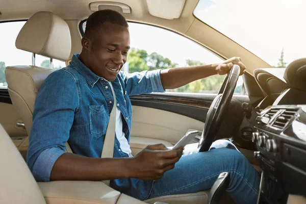 Sonriente chico negro usando teléfono inteligente mientras conduce el coche —  Fotos de Stock
