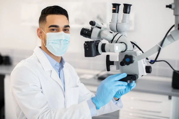 Árabe dentista masculino vestindo máscara médica protetora usando microscópio dentário na clínica — Fotografia de Stock