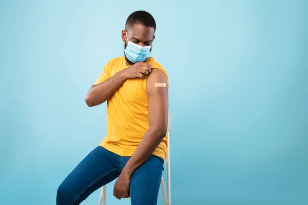 Ragazzo afroamericano in maschera viso mostrando la spalla con benda adesiva dopo la vaccinazione covid-19, sfondo blu — Foto Stock