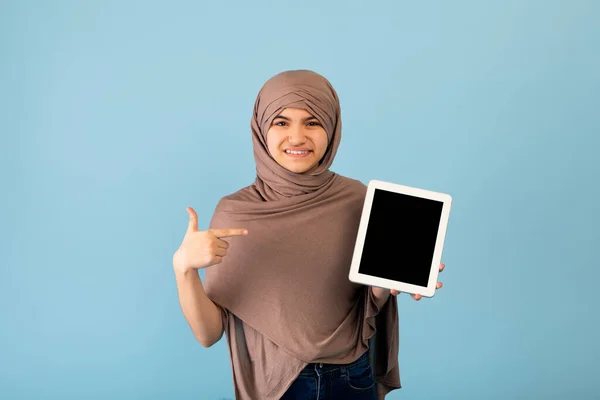 Gadis remaja yang gembira dalam jilbab Muslim menunjuk pada tablet pc dengan layar kosong di latar belakang biru, mockup untuk aplikasi atau situs web — Stok Foto