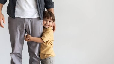 Küçük mutlu çocuk babasına sarılıyor, kameraya gülümsüyor, sevildiğini ve güvende olduğunu hissediyor, gri duvarın üzerinde boş bir alanla poz veriyor.