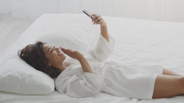 Wanita muda afrika yang bahagia berbaring di tempat tidur dan konferensi video dengan keluarga melalui ponsel, beristirahat di kamar hotel — Stok Video