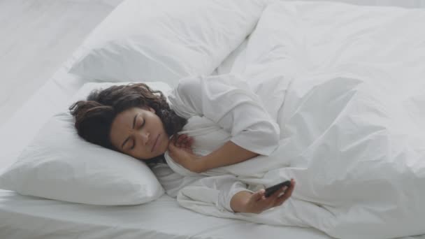 Δυσάρεστη Αφροαμερικανή κυρία που κλείνει το ξυπνητήρι στο κινητό, γυρίζει και κοιμάται στο κρεβάτι. — Αρχείο Βίντεο