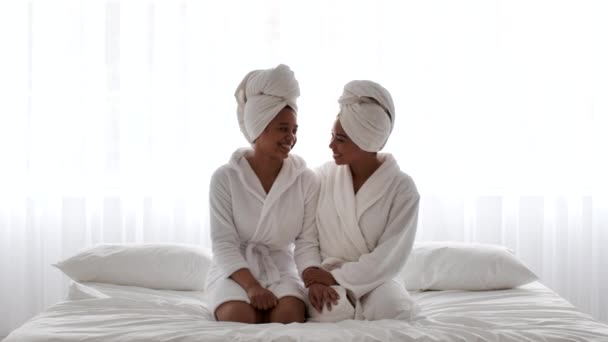 Thuis Spa. Twee lachende Afro-Amerikaanse zusters dragen badjassen zittend op bed — Stockvideo