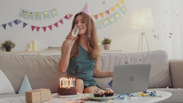 Feliz fiesta en línea. Joven alegre usando gorra de cumpleaños volando galleta y bebiendo champán bajo confeti — Vídeos de Stock
