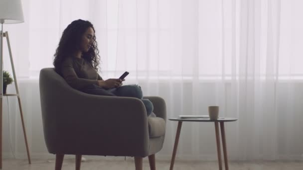 Mladá mírumilovná žena sedí v pohodlném křesle doma a píše smartphone, on-line chatování s přáteli — Stock video