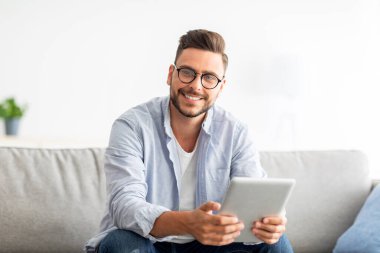 Teknoloji konsepti. Heyecanlı milenyum adamı dijital tablet kullanıyor, modern dairesindeki koltukta oturuyor.