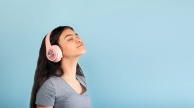 Kablosuz kulaklık takan sakin Hintli genç kız mavi arka planda kapalı gözlerle müzik dinliyor, boşluğu kopyalıyor.