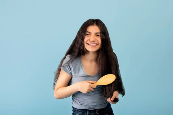 Brunette jeune fille indienne brossant ses beaux cheveux longs avec une brosse en bois naturelle sur fond de studio bleu — Photo