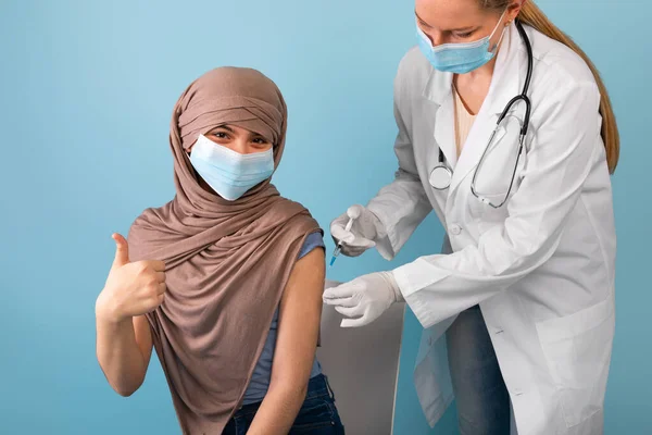 Muchacha adolescente musulmana en hiyab y máscara médica recibiendo covid vacuna inyección de médico femenino, gesto pulgar hacia arriba —  Fotos de Stock