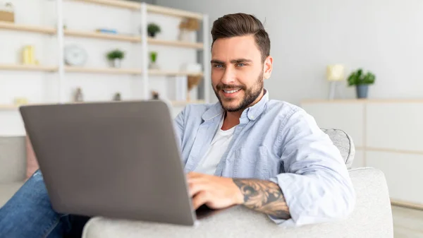 Tipo positivo reclinado en el sofá con el ordenador portátil en el interior del hogar, navegar por Internet o desplazarse por las redes sociales, panorama — Foto de Stock