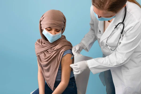 Muchacha adolescente musulmana en hijab usando mascarilla facial, recibiendo la vacuna contra el coronavirus de una doctora sobre fondo azul — Foto de Stock