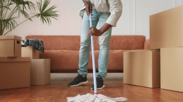 Καθαρισμός μετεγκατάστασης. Αγνώριστος Αφρικανός Αμερικανός άνδρας που πλένει το πάτωμα με σφουγγαρίστρα σε νέο διαμέρισμα ανάμεσα σε κινούμενα κουτιά — Αρχείο Βίντεο