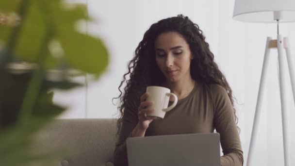 Jongedame digitale nomade werken online vanuit huis, typen op laptop en het drinken van hete koffie uit kopje, tracking shot — Stockvideo