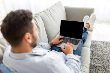 Reklam konsepti. Genç adam boş ekranlı modern dizüstü bilgisayar kullanıyor, koltukta oturuyor, model alıyor, omzunun üstünden çekim yapıyor.