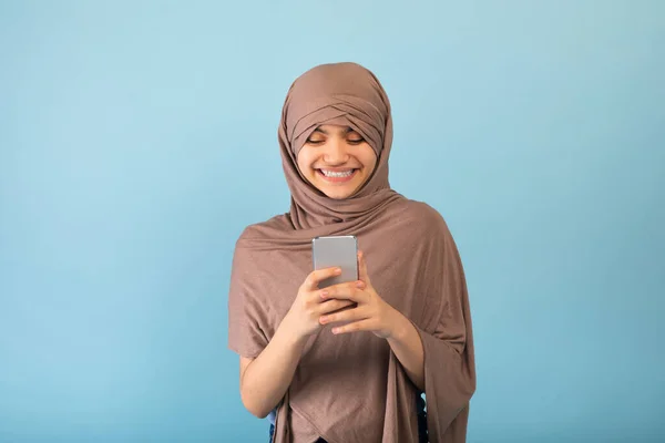 Gadis remaja Muslim positif mengenakan jilbab menggunakan ponsel, memeriksa pesan, menjelajah media sosial melalui latar belakang studio biru — Stok Foto