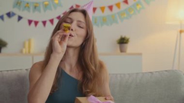 Doğum günün kutlu olsun. Bayram şapkası takan, ıslık çalan ve kameraya hediye kutusu sunan heyecanlı genç bayan.