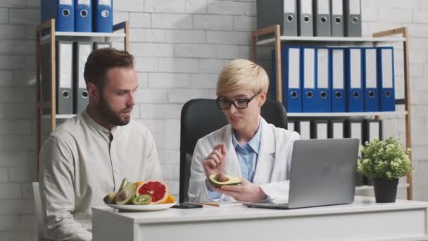 Nutricionista positiva hablando con paciente masculino, explicando los beneficios de la grasa vegetal, mostrando aguacate — Vídeo de stock