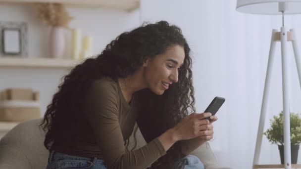 Buenas noticias. Joven mujer rizada positiva surfeando en las redes sociales en el teléfono móvil, disfrutando de noticias increíbles, sentado en el sillón — Vídeo de stock