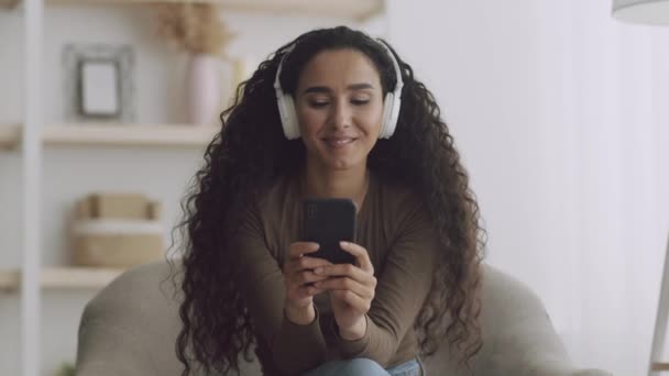 Relax tijd. Jonge krullende dame thuis, luisteren naar muziek in koptelefoon en netwerken op mobiele telefoon — Stockvideo