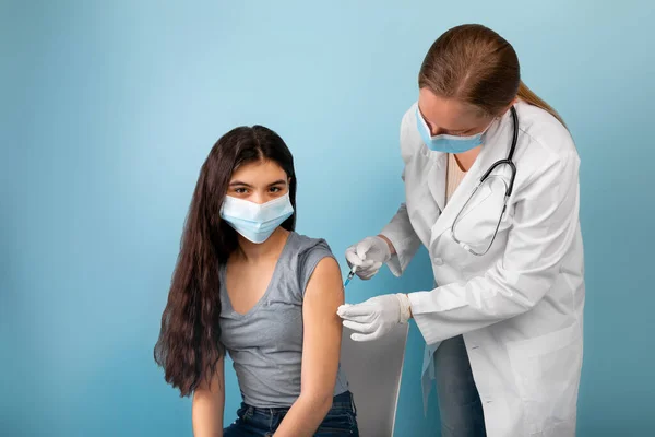 Médico haciendo la inyección de la vacuna contra el coronavirus a la adolescente india en la máscara facial en fondo de estudio azul — Foto de Stock