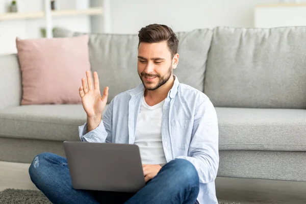 Szczęśliwy człowiek millennial machając ręką do laptopa wideo rozmowy siedząc na podłodze we wnętrzu salonu, wolne miejsce — Zdjęcie stockowe