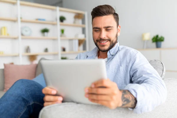 Fechar-se do homem caucasiano descansando no sofá com tablet digital, usando aplicativo móvel agradável, interior da casa, espaço livre — Fotografia de Stock