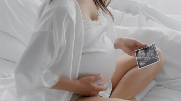 Mujer embarazada irreconocible sosteniendo foto del ultrasonido del bebé y tocando el vientre en la cama — Foto de Stock