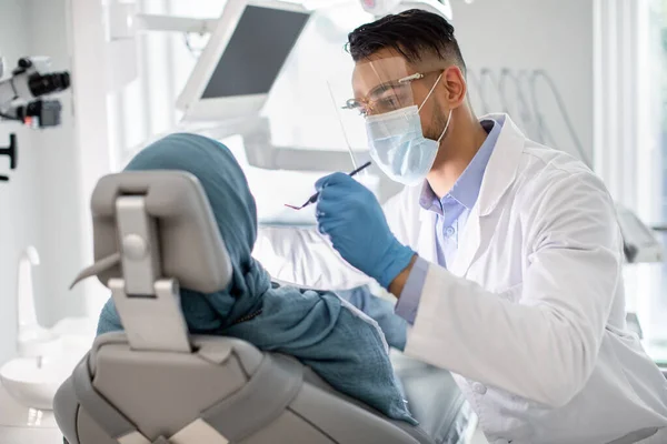 Dentista masculino de Oriente Medio en máscara médica y escudo facial revisando los dientes de los pacientes — Foto de Stock