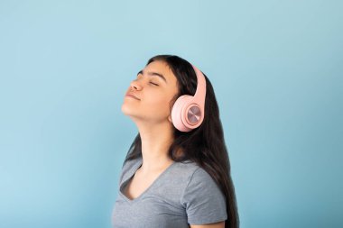 Çekici Hintli genç kız yeni stereo kulaklığının keyfini çıkarıyor, mavi arka planda kapalı gözlerle müzik dinliyor.