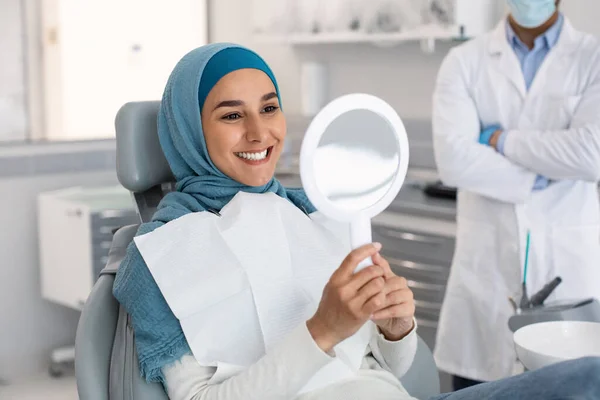 Paciente muçulmana feliz olhando para o espelho após o tratamento na clínica dentária — Fotografia de Stock