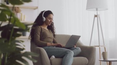Kulaklıklı olumlu genç bayan favori müzik beyazı evde dizüstü bilgisayarla çalışıyor.