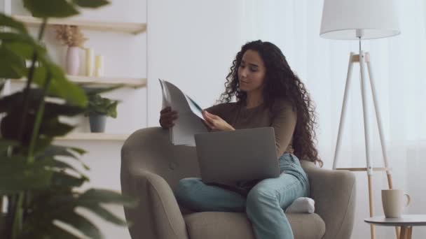 Νεαρή σγουρή γυναίκα που εργάζεται στο σπίτι, διαβάζει έγγραφα και συγκρίνει πληροφορίες με φορητό υπολογιστή, κάθεται άνετα στην πολυθρόνα — Αρχείο Βίντεο