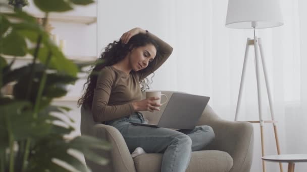 Slappna av på helgerna. Ung fridfull dam med kopp kaffe sittande i fåtölj och titta på film online på bärbar dator — Stockvideo